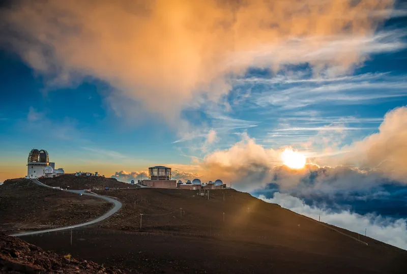 Dramatic sunrise at Haleakala summit showing the moment the sun breaks over the horizon, illuminating a vast sea of clouds below the volcanic crater rim, with silhouettes of visitors in winter clothing watching in awe