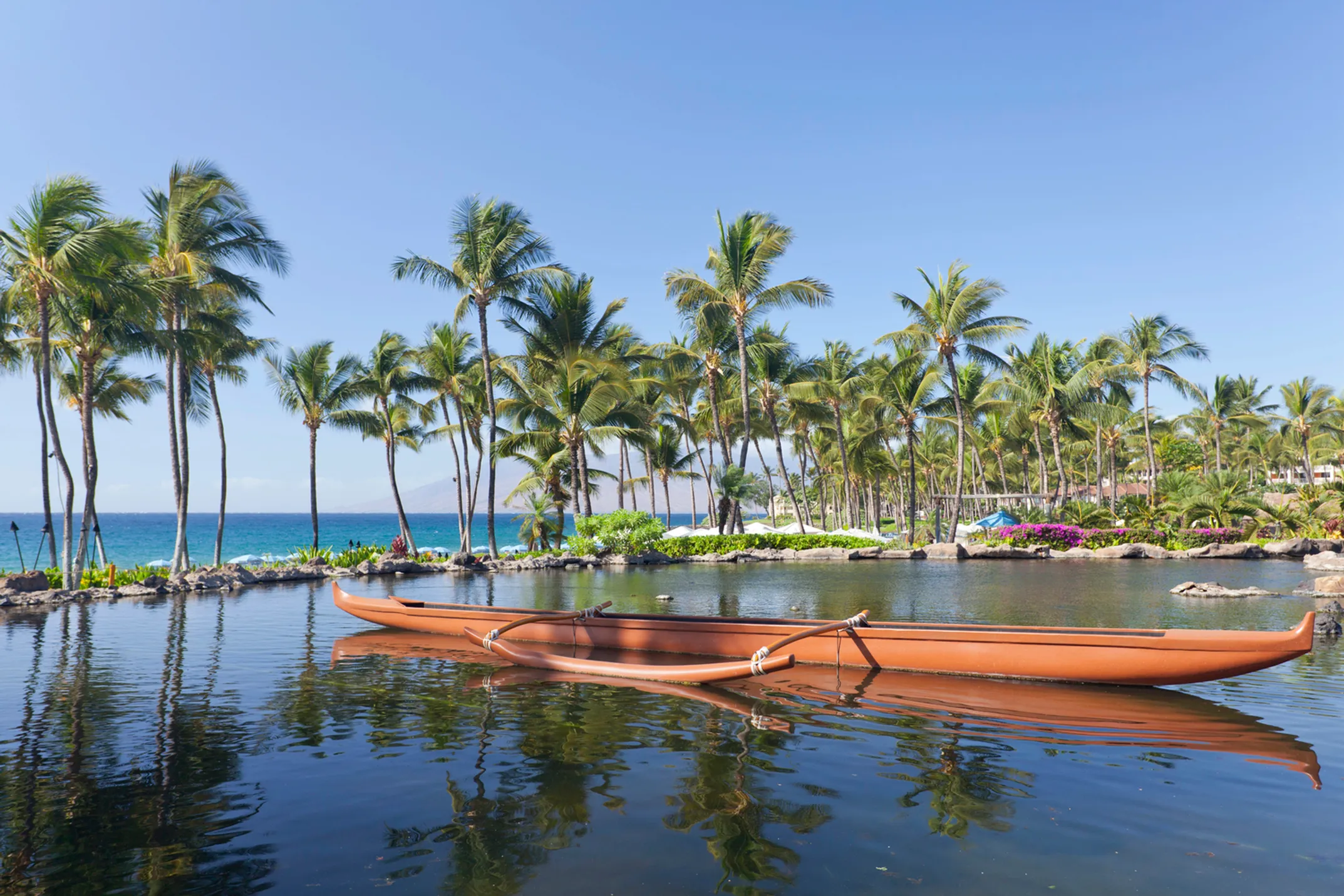 Traditional Hawaiian outrigger canoe crafted from koa wood with six paddlers in action, racing across blue Hawaiian waters with mountains in distance