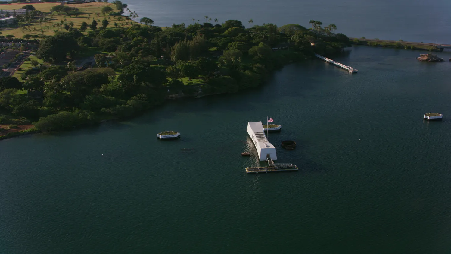 Aerial view of Pearl Harbor with the USS Arizona Memorial visible, mountains in the background, and modern Honolulu skyline in the distance