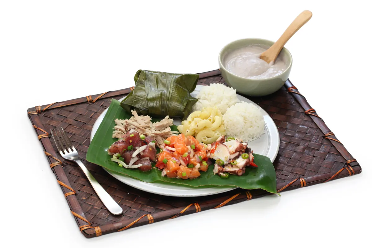Traditional Hawaiian foods including poke, kalua pig, rice, and poi served on banana leaves with traditional woven mat