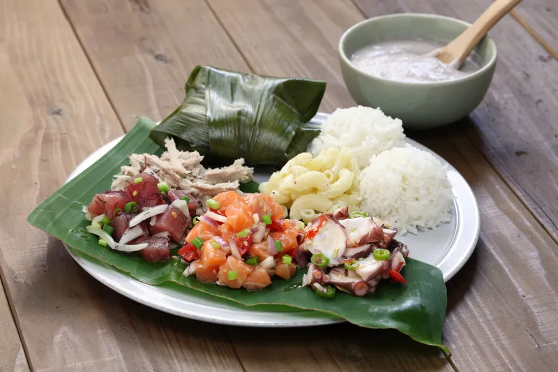Traditional Hawaiian ohana (family) sharing meal with poi, kalua pig, and other local dishes on lauhala mats with scenic island backdrop